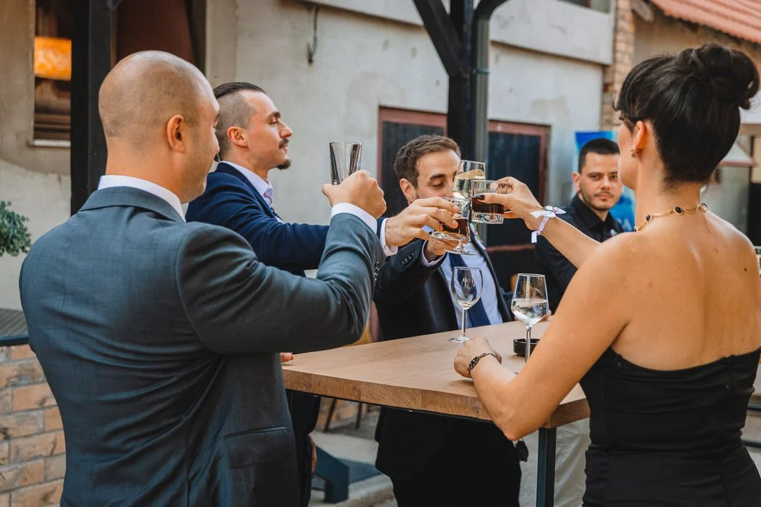 Five adults at an outdoor gathering, clinking glasses in a toast near a wooden table.