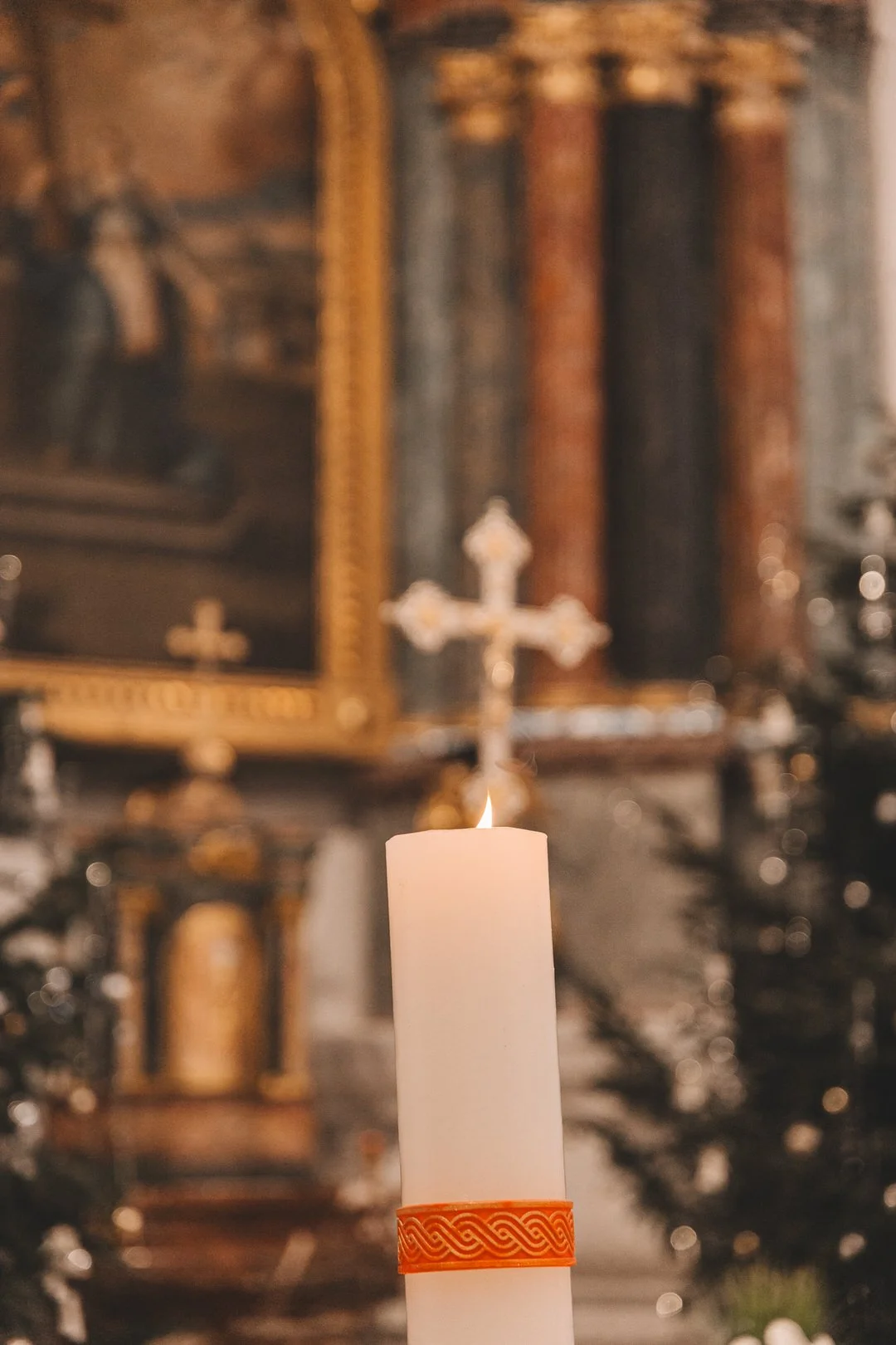 A lit candle with an orange decorative band around its base, inside a church  with a crucifix and religious paintings in the background.