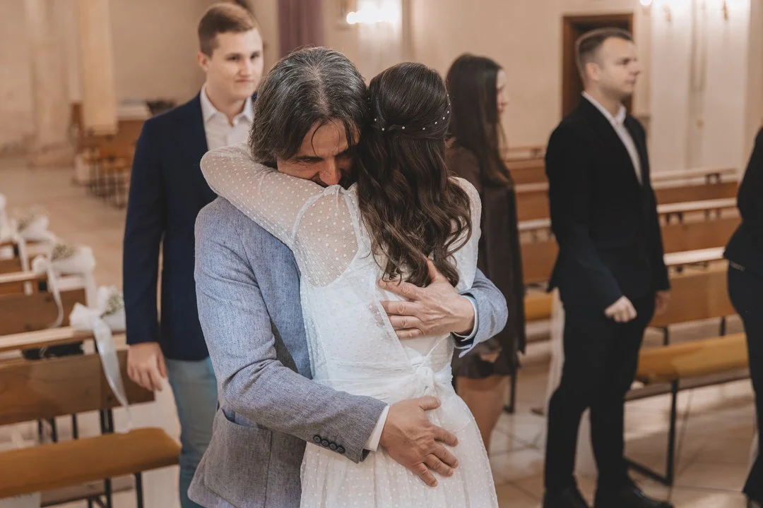 A bride and her father embrace in a hug at a wedding ceremony inside a church, with other guests standing in the background.