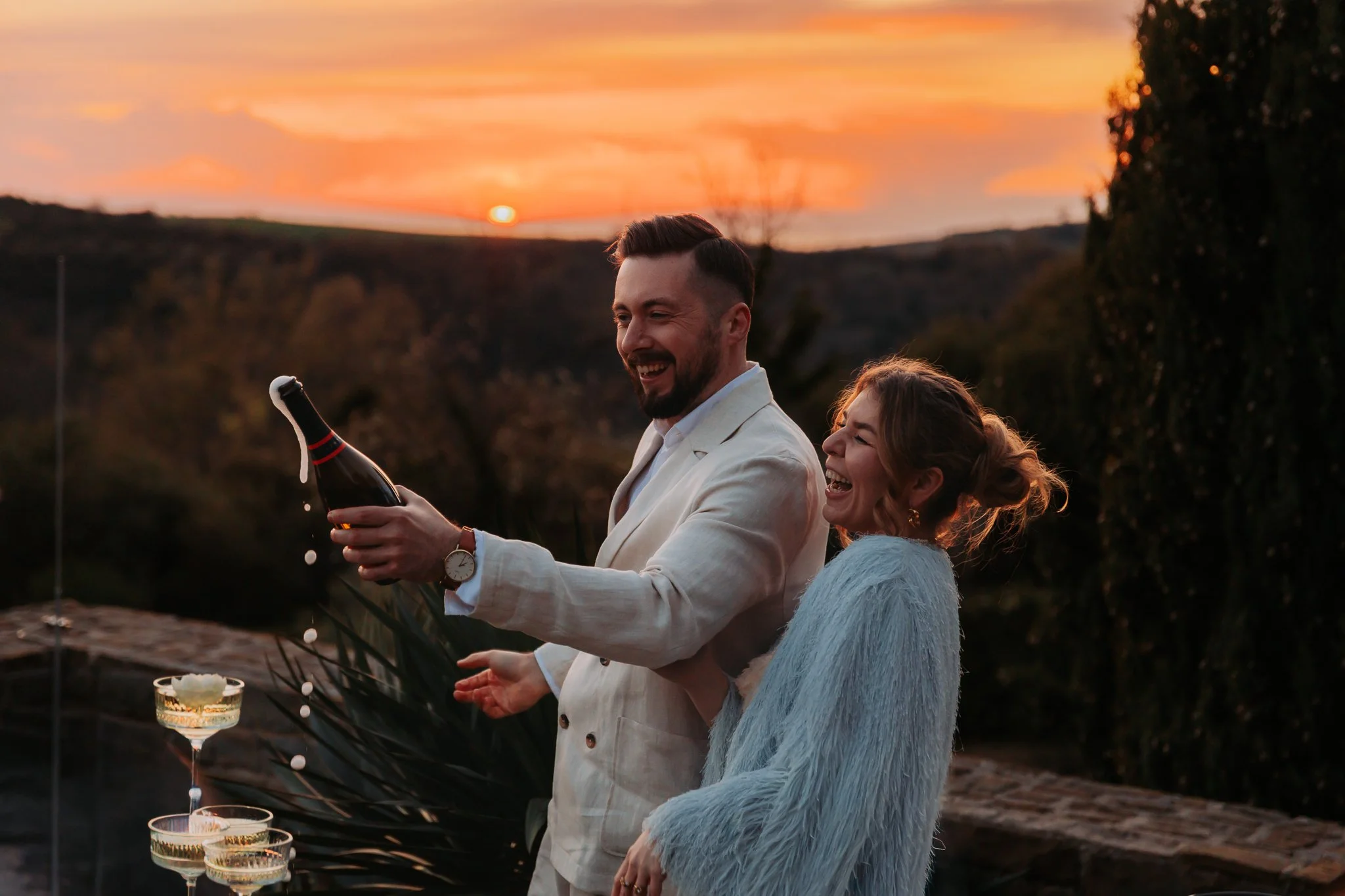 A man in a white suit and a woman in a light blue dress celebrating outdoors at sunset with champagne and candlelight.