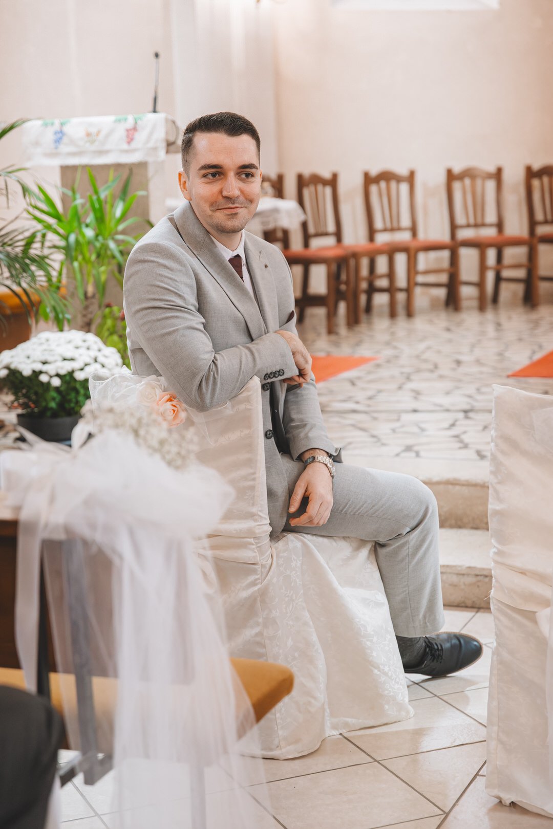 A groomsman in a light gray suit sitting on a decorated chair at a formal event, with chairs and a floral arrangement around him.