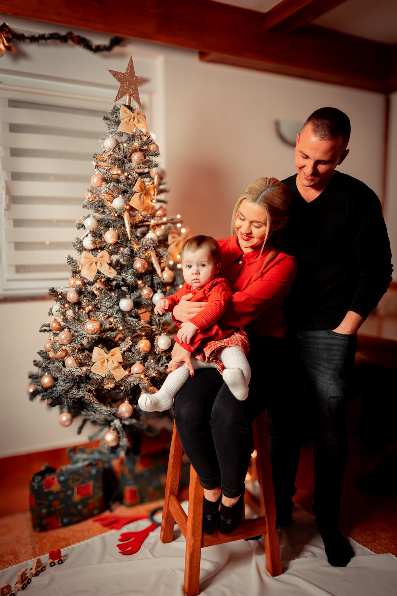 A family celebrating Christmas with a decorated tree, a woman holding a young girl on a ladder, and a man standing beside them. Gift boxes are underneath the tree, and holiday decorations are in the background.