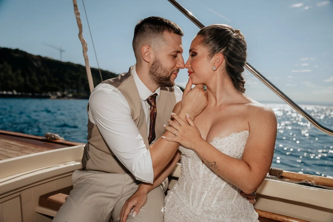 Couple on a boat, in wedding attire, with their foreheads touching, holding hands, on sunny day with ocean background.
