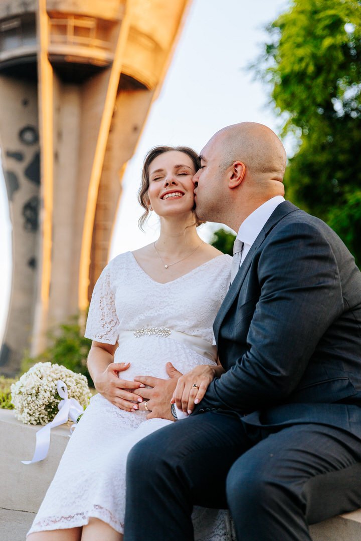 A man kissing a pregnant woman on the cheek at an outdoor wedding ceremony under a Vukovar water towe, with a bouquet of white flowers nearby.