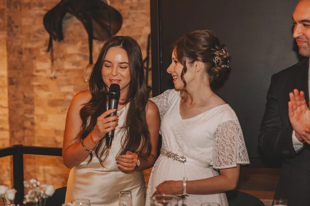 A woman in a white dress giving a speech with a microphone, standing next to a woman in a wedding dress, with a man clapping nearby in a celebration setting.