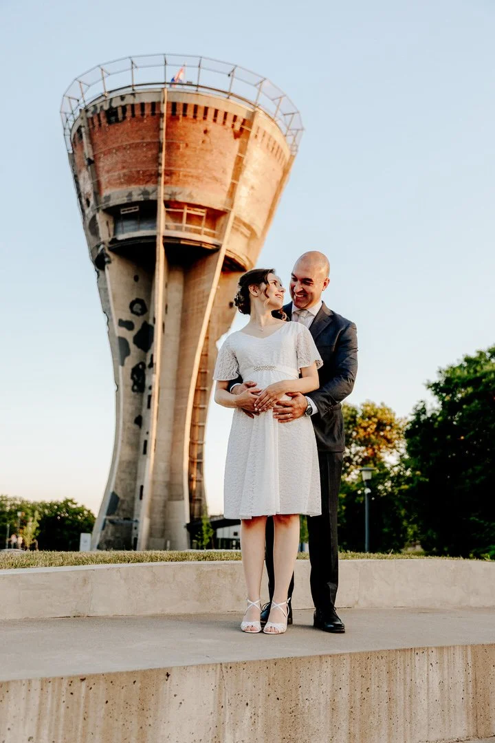 Bride and groom standing outdoors at sunset, with the iconic Vukovar Water Tower in the background, during their wedding photoshoot in Vukovar, Croatia.