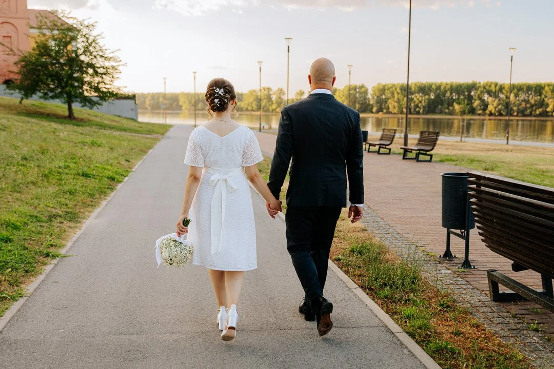 A bride and groom walking hand-in-hand along a park pathway near a Dunav river, with benches and lampposts, during early evening or late afternoon.