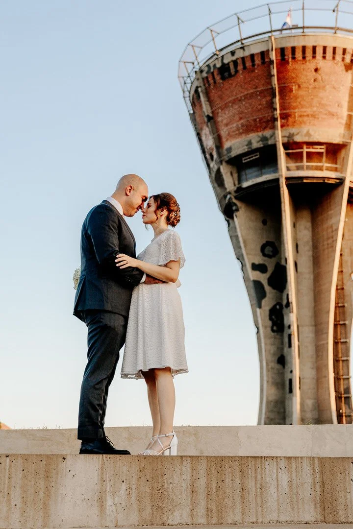 A couple dressed in formal attire standing close, forehead to forehead, outdoors during sunset, with an old watertower in  Vukovar in the background.