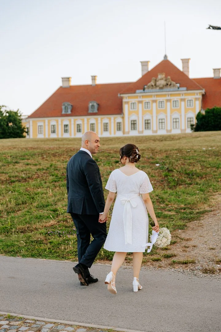 A newlywed couple holding hands and walking along a paved path with Eltz Castle in Vukovar, Croatia, in the background during daytime.