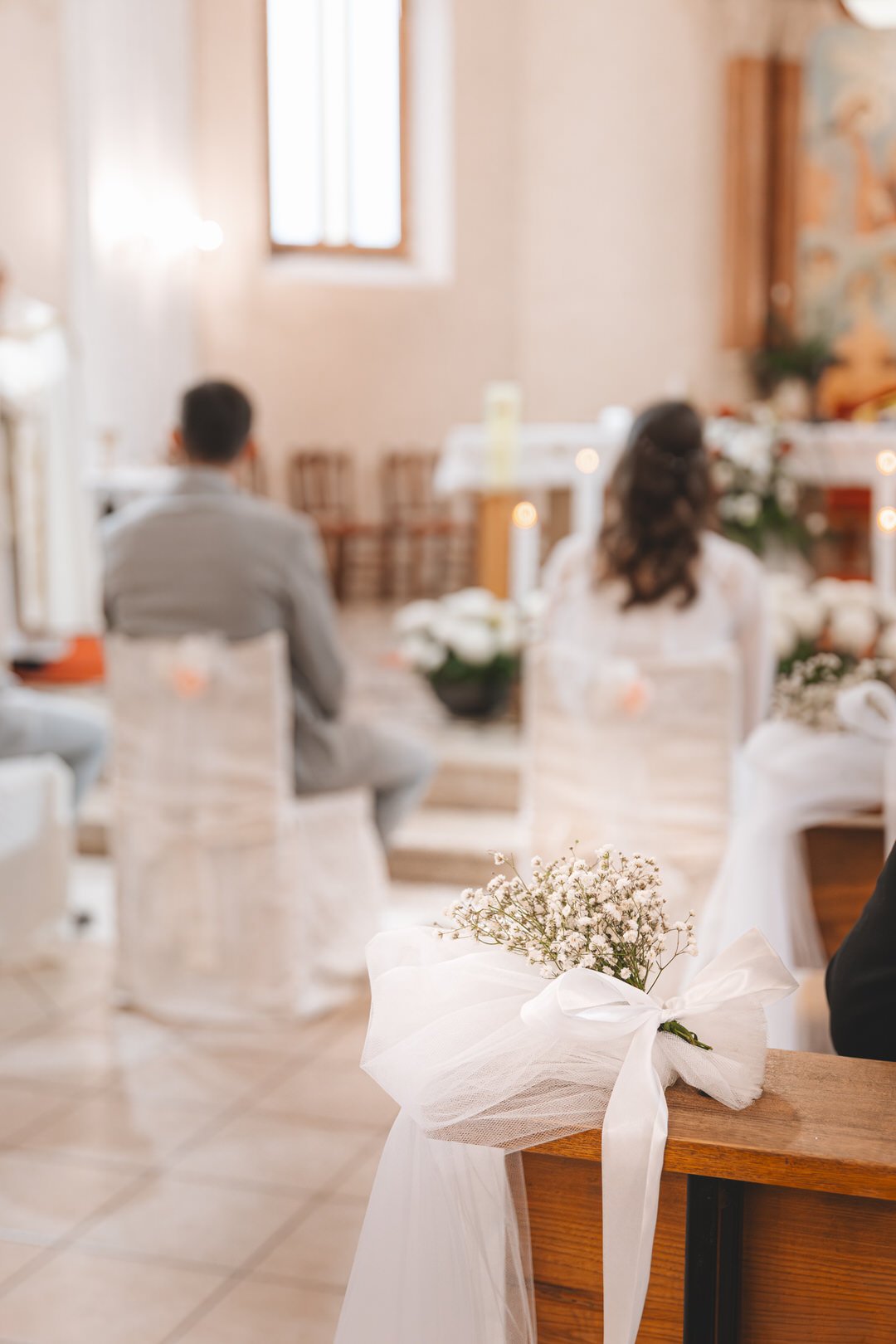 A bouquet of small white flowers wrapped with white tulle and ribbon on a wooden pew in a church interior, with bride and groom sitting in the background during a wedding ceremony.