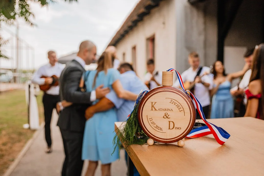 A wooden wedding favor with engraved names and date, decorated with greenery and ribbons, on a table at a wedding celebration with guests dancing and playing guitars in the background.