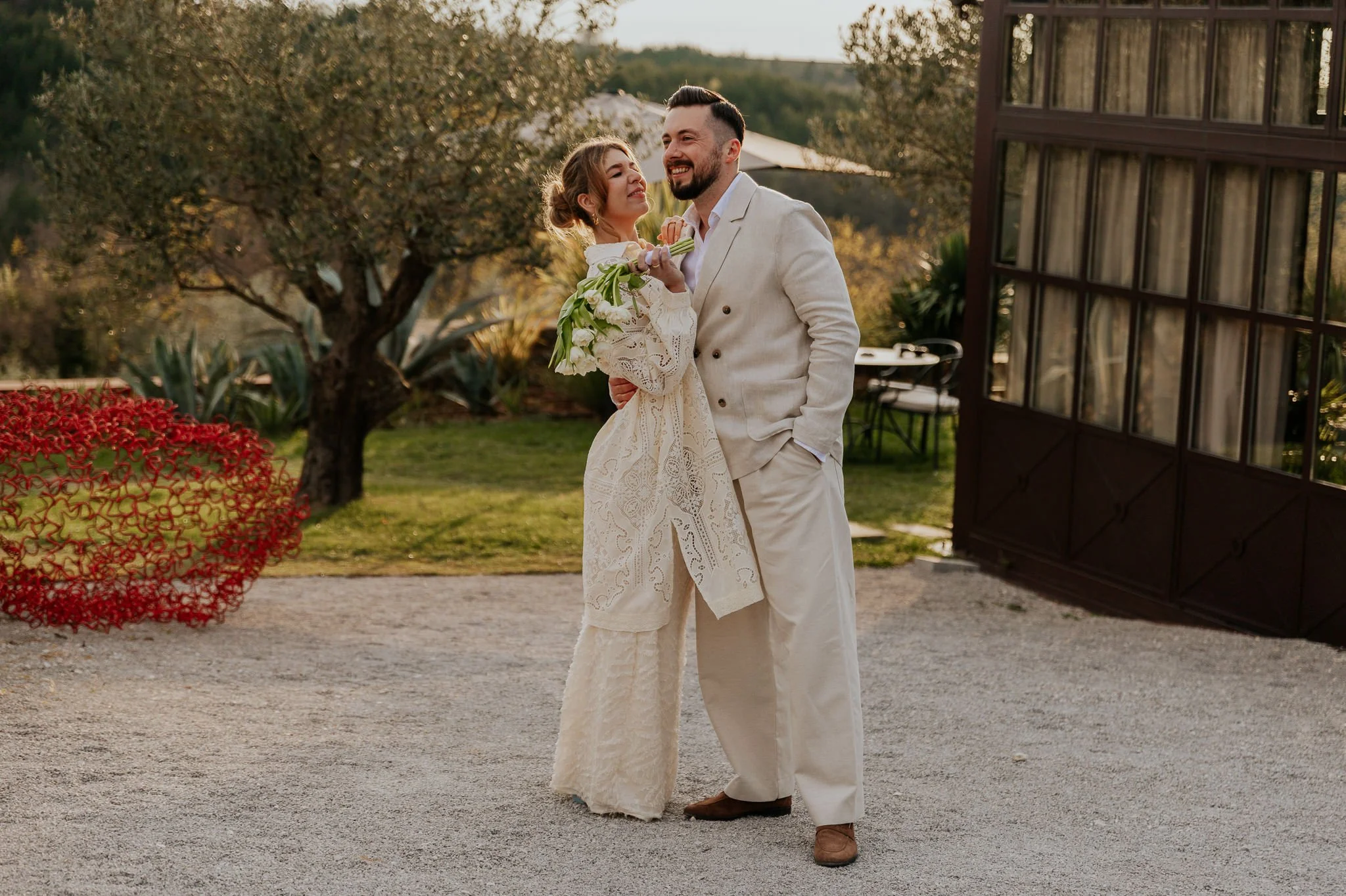 A couple dressed in wedding attire smiling and looking at each other outdoors during sunset, with trees and a building with large windows in the background at San Canzian Istria, Croatia