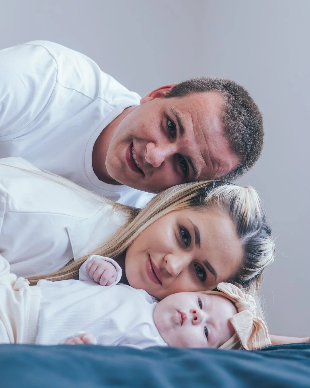 A family portrait of three people, a man, a woman, and a baby, lying down together. They are all smiling and dressed in white. The man is at the top, the woman in the middle, and the baby at the bottom, with the baby wearing a headband.