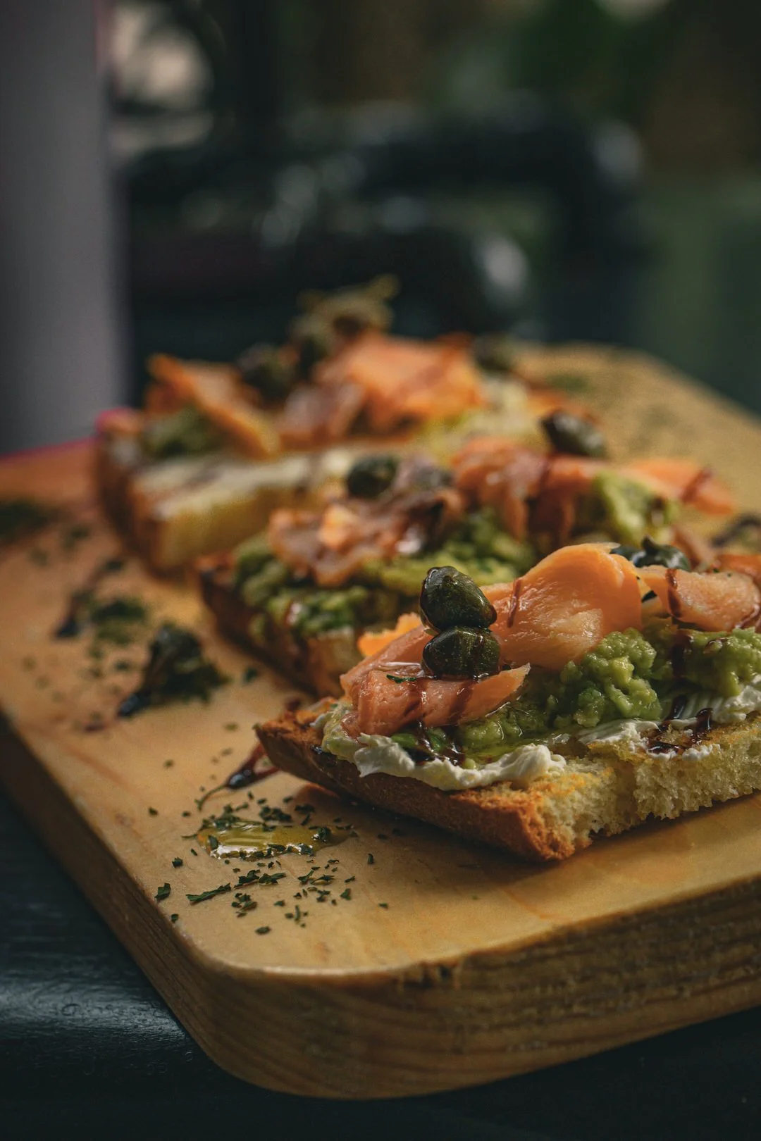 Close-up of three slices of toast with avocado, smoked salmon, black caviar, and herbs on a wooden serving board.