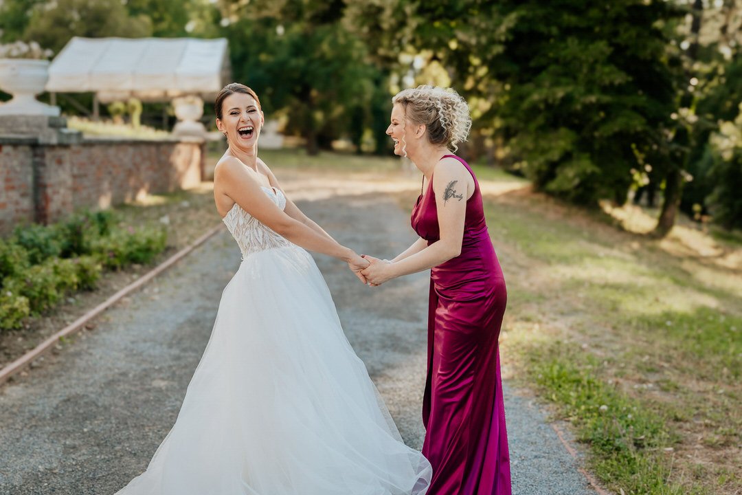 A bride in a white wedding dress and a maid of honor in a burgundy gown holding hands and laughing outdoors, with trees and a brick structure in the background.