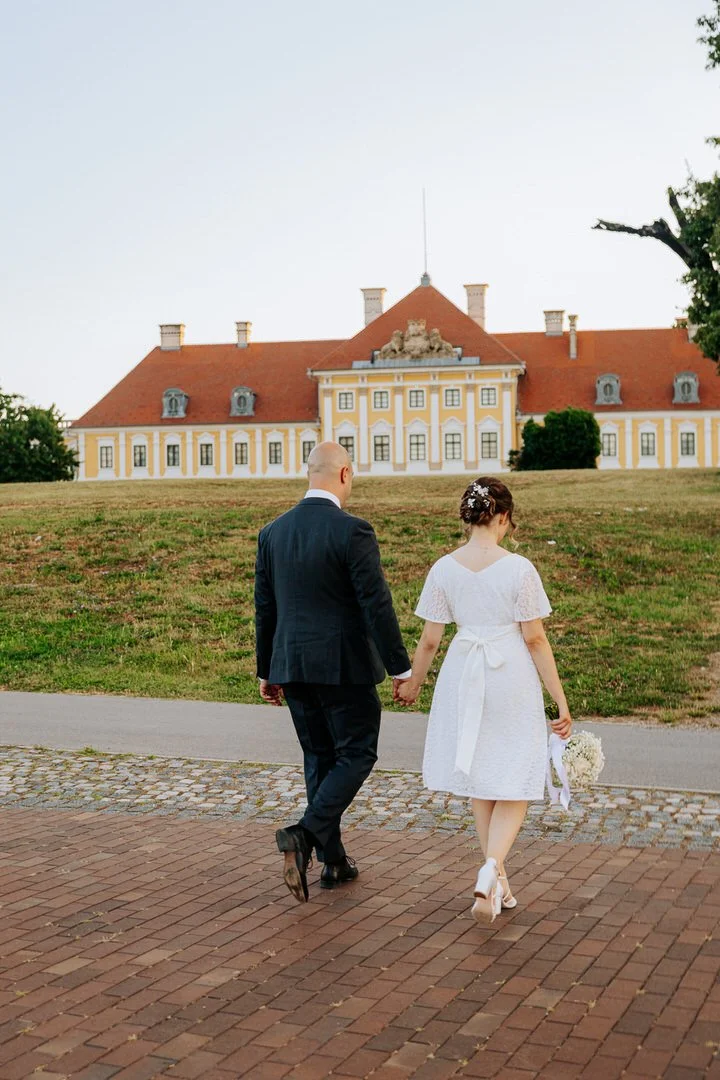 A newlywed couple holding hands and walking towards a large, historic yellow and white building with a red roof named Eltz in Vukovar