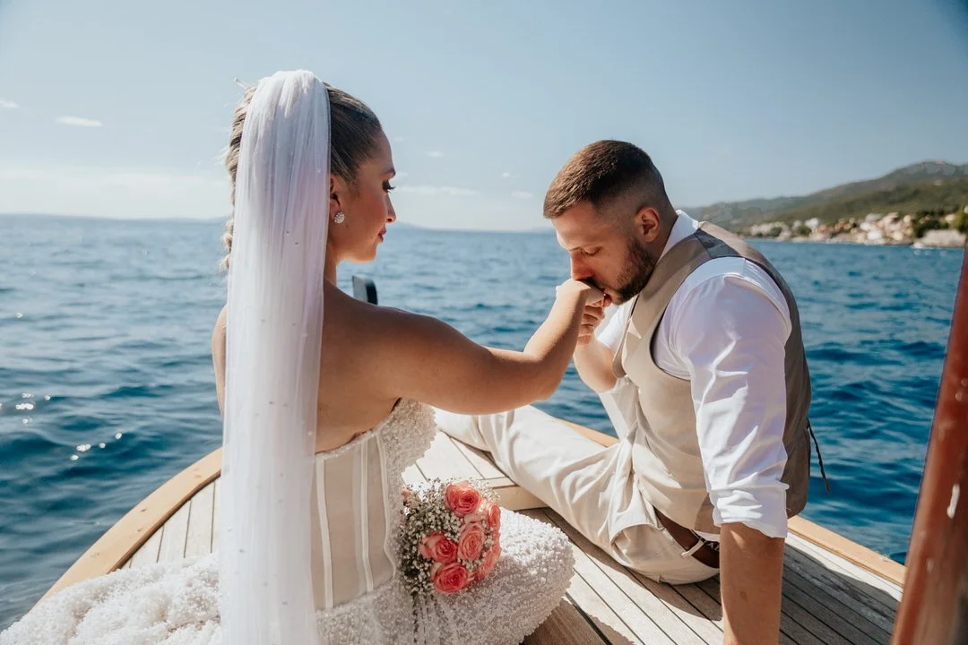 A bride and groom on a boat at sea; the groom kisses the bride's hand, and the bride is holding a bouquet of pink roses.