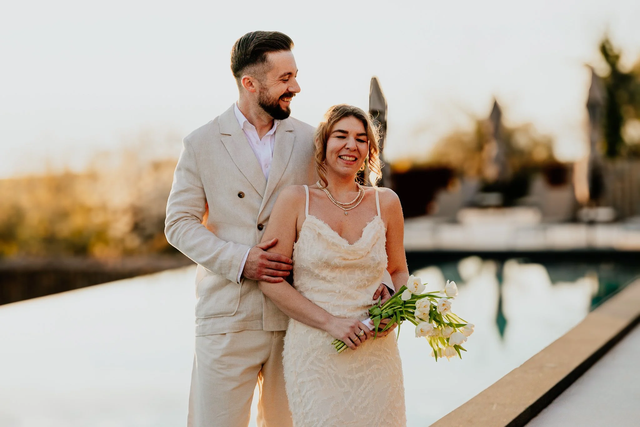 A newlywed couple standing outdoors near water at sunset, smiling and holding a bouquet of white roses, with the groom in a beige suit and the bride in a lace gown.