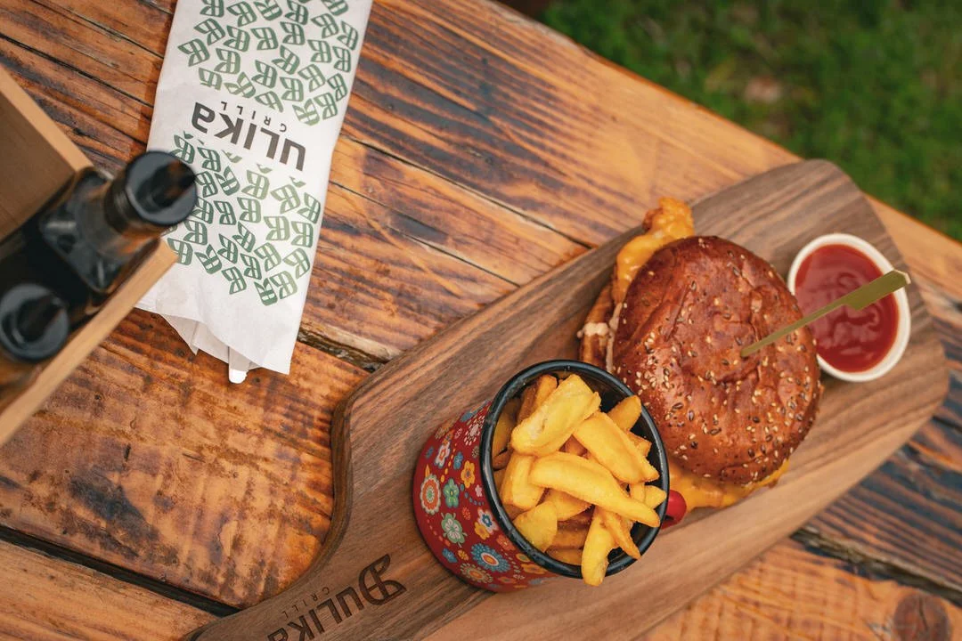 A cheeseburger with sesame seed bun, a side of French fries in a decorative cup, and ketchup in a small white container on a wooden serving board on a outdoor wooden table.
