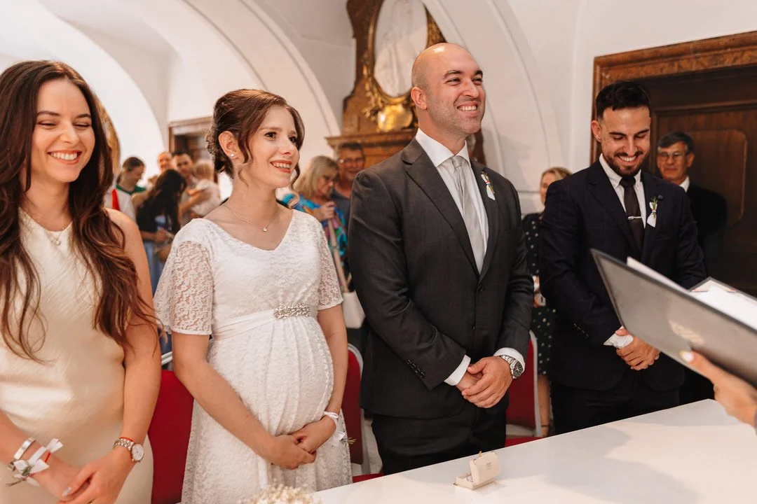 People standing in a line at a wedding ceremony, including a pregnant woman, three men in suits, and onlookers in the background.