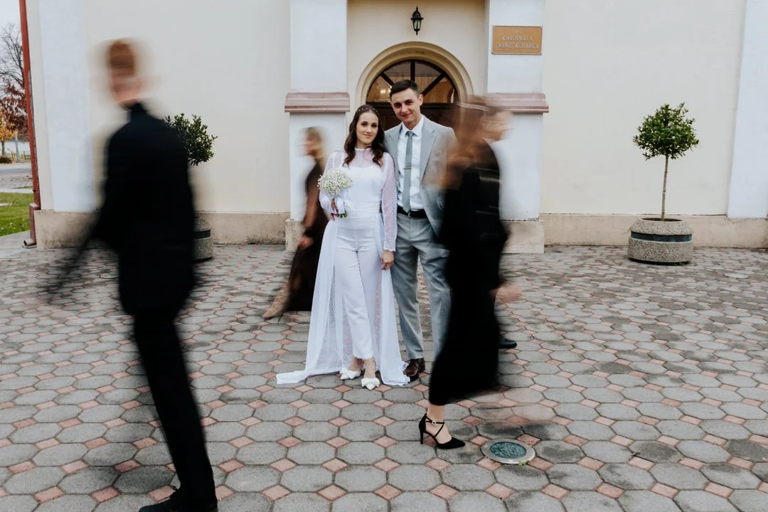 A newlywed couple standing in front of a church, surrounded by blurred walking guests.