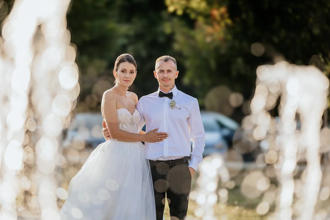 Bride and groom standing outdoors, wedding attire, trees in the background, sunlight, romantic setting.
