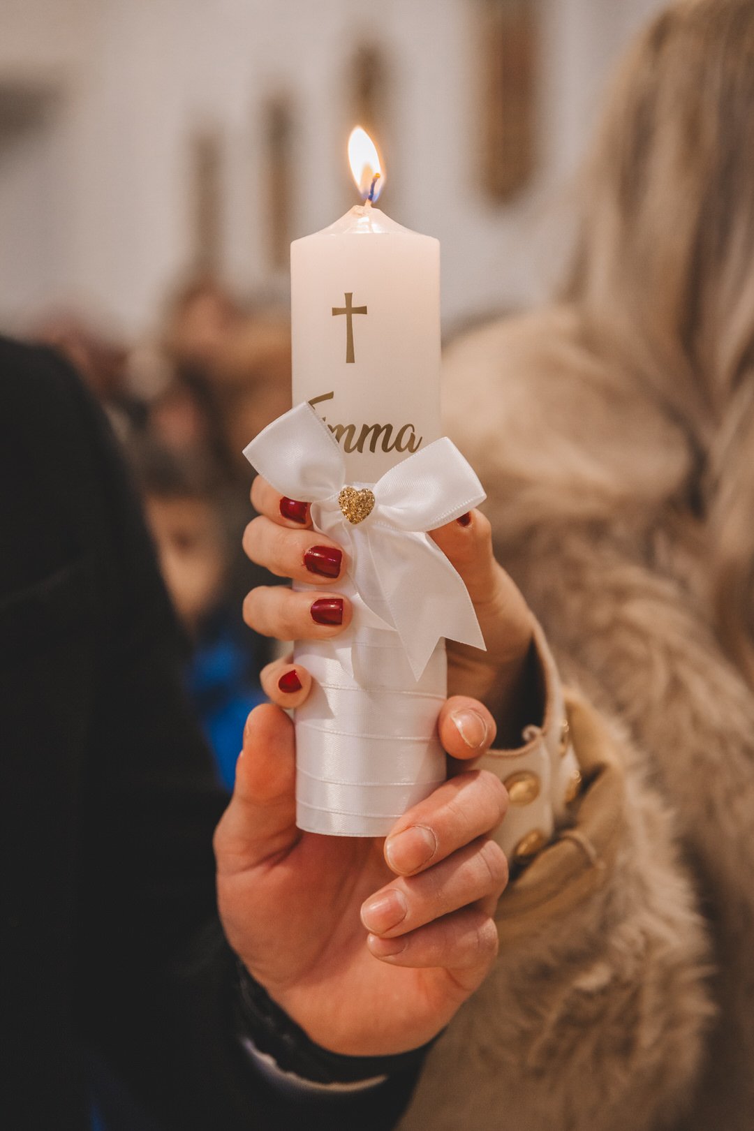 Person holding a white candle decorated with a white ribbon bow and a gold heart with the name Emma written on it, in a church setting.