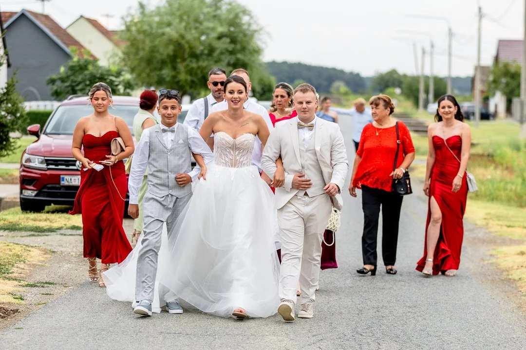 A bride in a white wedding gown and a groom in a white suit walking down a street with friends and family.