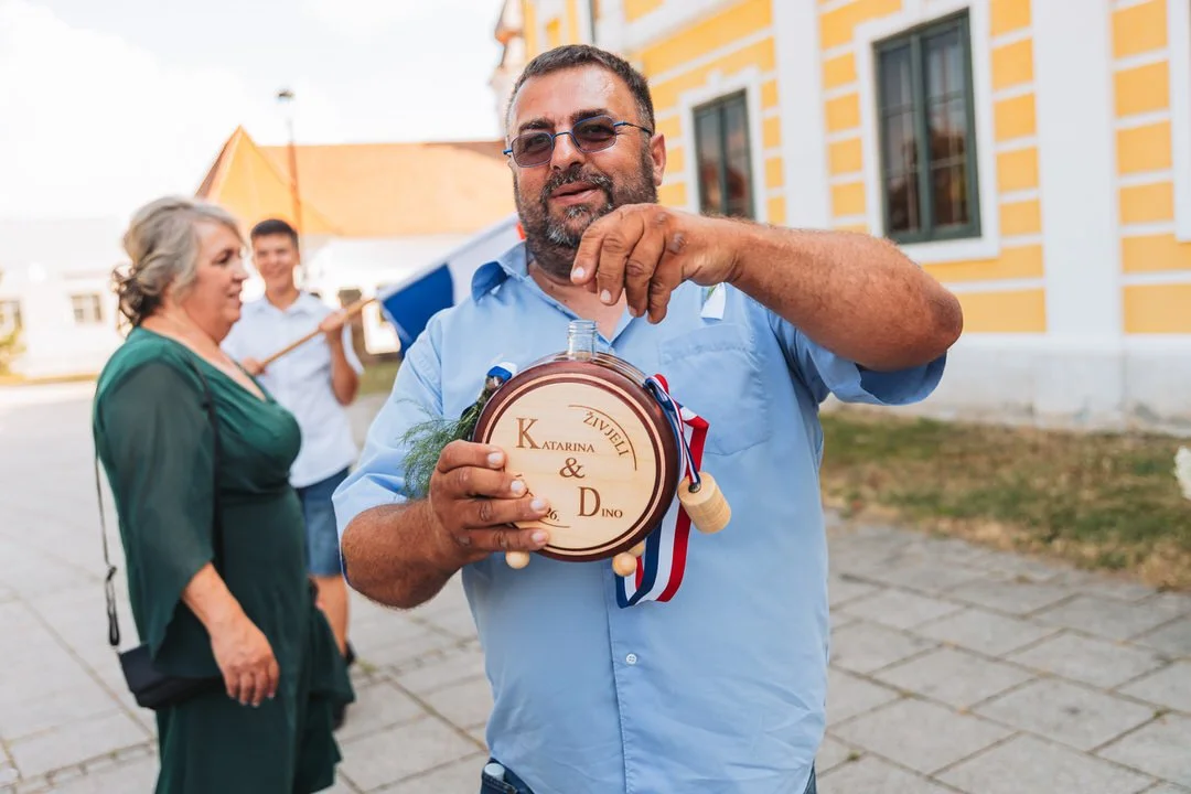 Man holding a personalized wooden flask with a cork, ribbon, and greenery, outdoors in front of a yellow building, with two women and one man in the background.