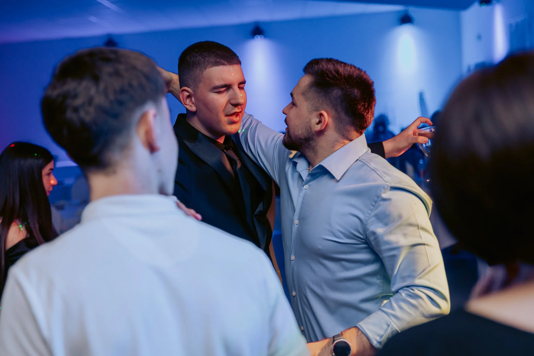 Two men are dancing closely at a party with blue lighting. One man in a black shirt and tie has his arm around the other, who is wearing a white shirt. Several other people are visible around them.