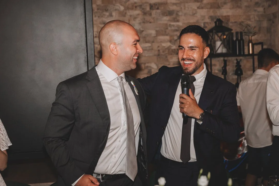 Two men in formal attire smiling and talking, one holding a microphone, at a social event or party in a room with a brick wall background.