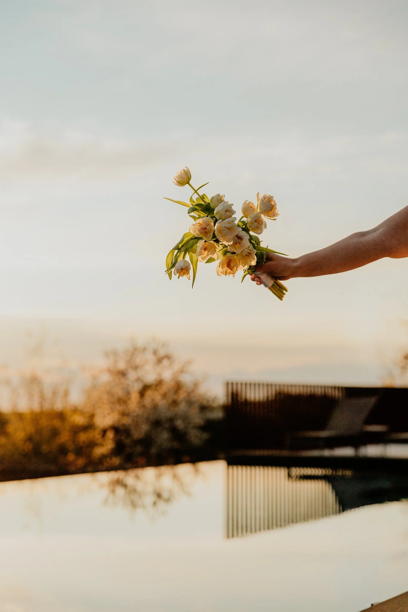 A person with a dark skin tone holding a bouquet of pale yellow and white flowers against a cloudy sky during sunset at a San Canzian wedding in Istria, Croatia.