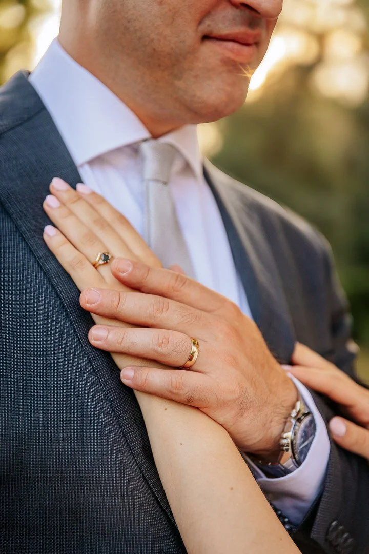 Close-up of a man and woman holding hands, with the man's hand resting on his chest and the woman's hand on top, both wearing wedding rings.
