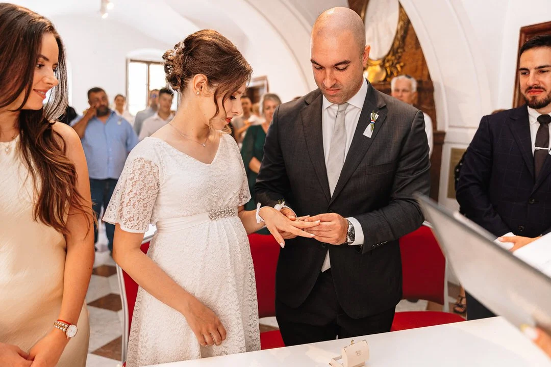 A couple getting married during a wedding ceremony, with a woman in a white dress and a man in a dark suit, exchanging rings with guests in the background.