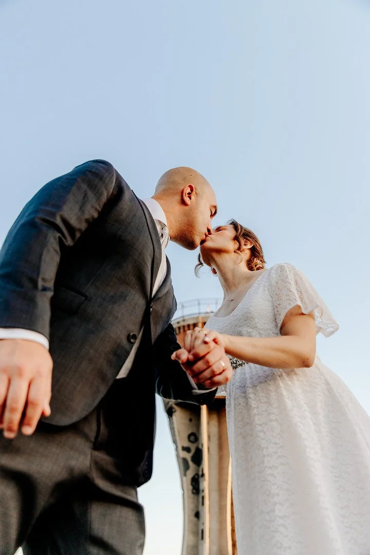 A couple in wedding attire sharing a kiss outdoors, with the sky in the background.