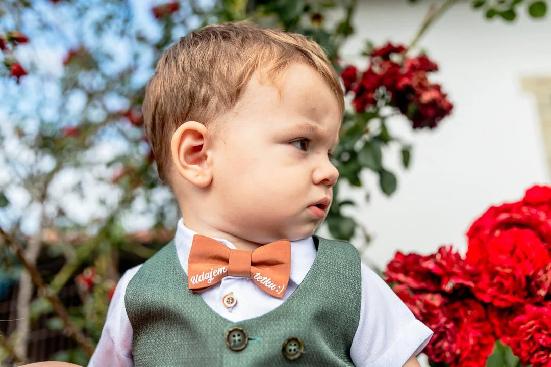 A young boy with red hair and fair skin standing outdoors with red flowers in the background, wearing a white shirt, a green vest, and a brown bow tie.