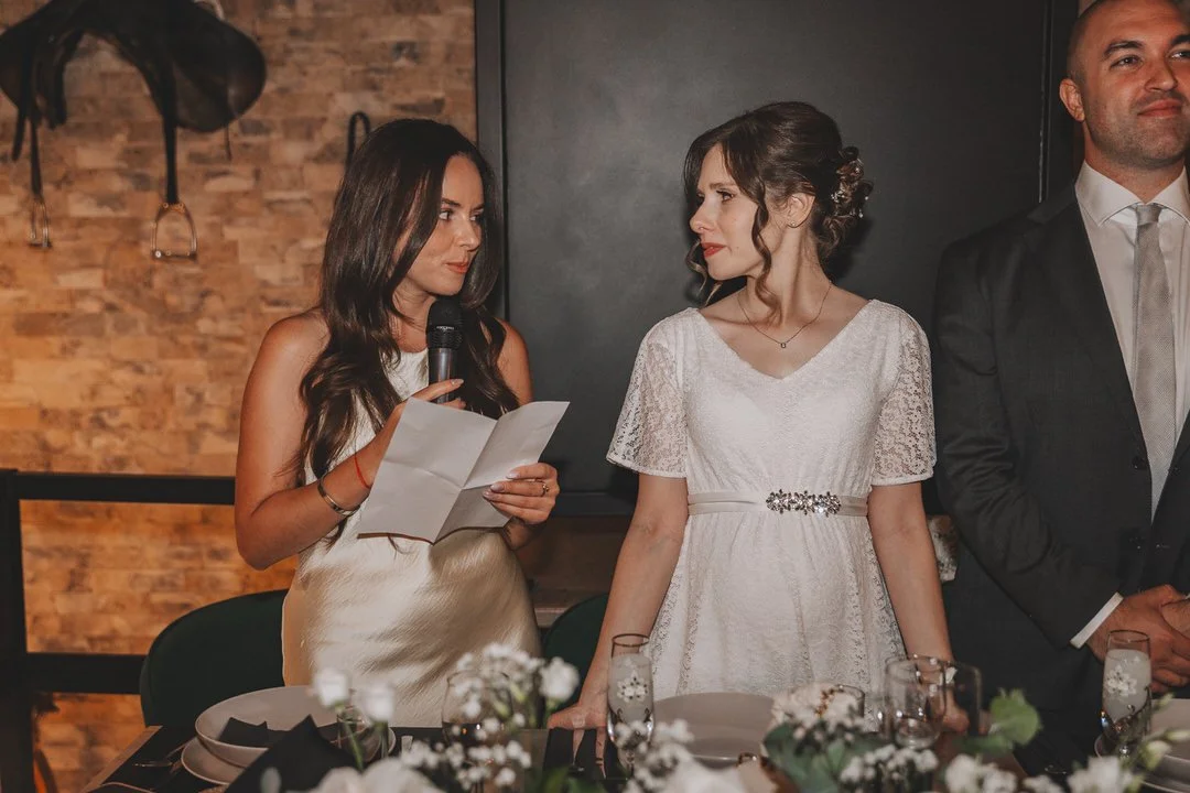 A woman in a beige dress holding a microphone and reading from a paper, standing next to another woman in a white wedding dress at a formal event.