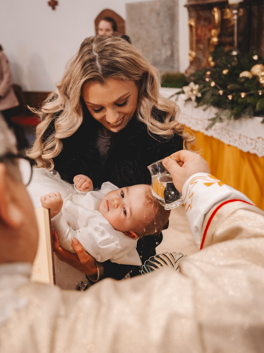 A woman holding a young girl during a Croatia baptism in Slavonija while a priest pours water on her head. green tree decorations are visible in the background.