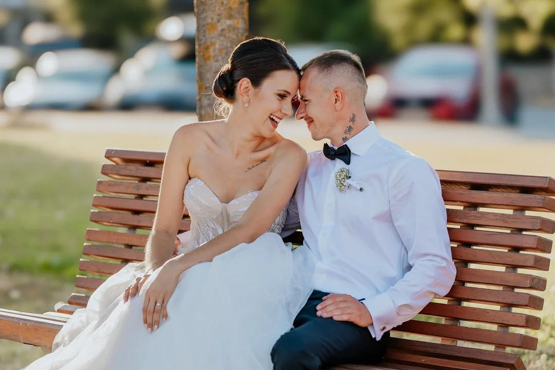 A bride and groom sitting close together on a wooden park bench, smiling and touching foreheads, with a background of trees and parked cars.