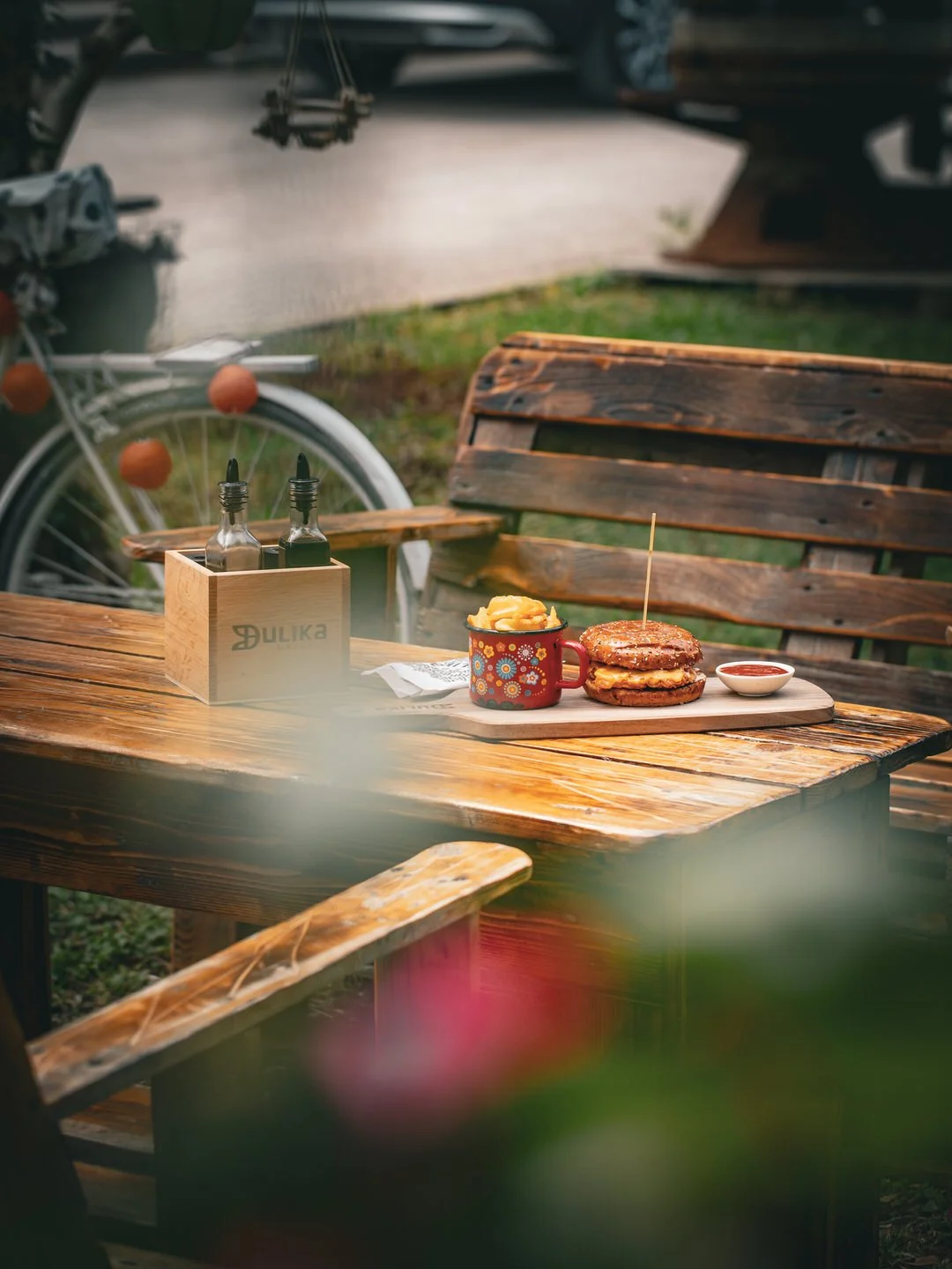 A wooden outdoor table with a burger, fries in a colorful mug, and a small bowl of ketchup. A wooden box with condiment bottles and a rustic wooden bench nearby. Blurred background with a bicycle and greenery.