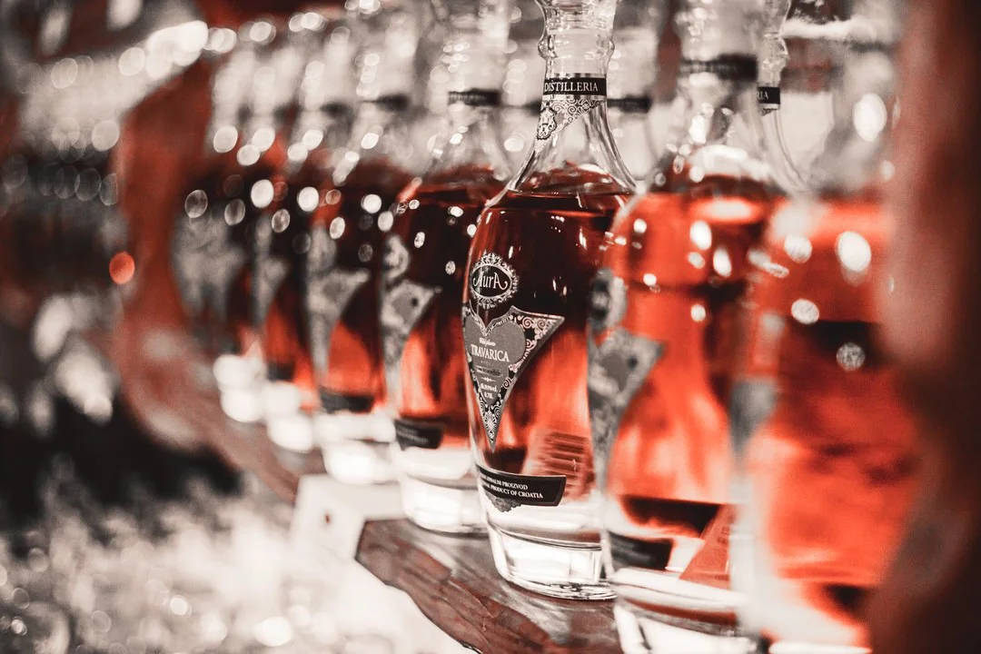 Multiple bottles of dark amber liquor arranged on a shelf, with focus on the nearest bottle in the center at Aura Buzet, Croatia.