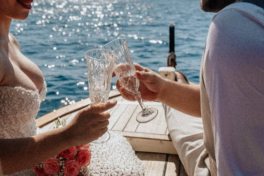 A woman in a wedding dress and a man in a white shirt toasting with champagne glasses on a boat in the water.