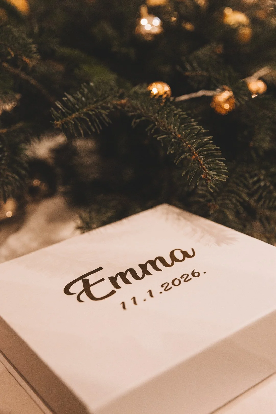 A white box with the name Emma and the date 11.1.2026 written on it, placed under a dark green Christmas tree with blurred lights in the background at a Croatia baptism in Slavonija.