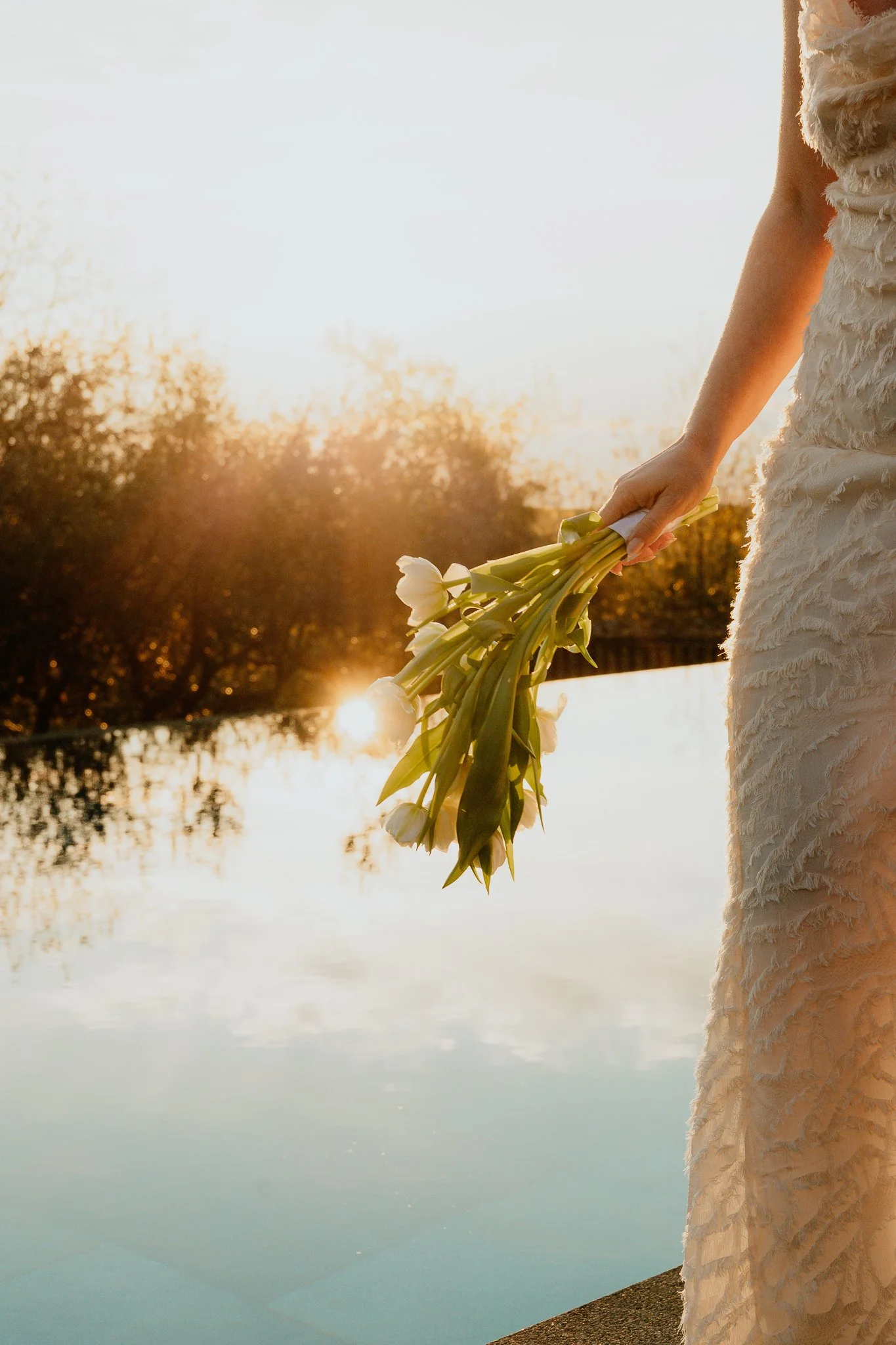 A person in a textured white dress holding a bouquet of white lilies at sunset near a body of water.
