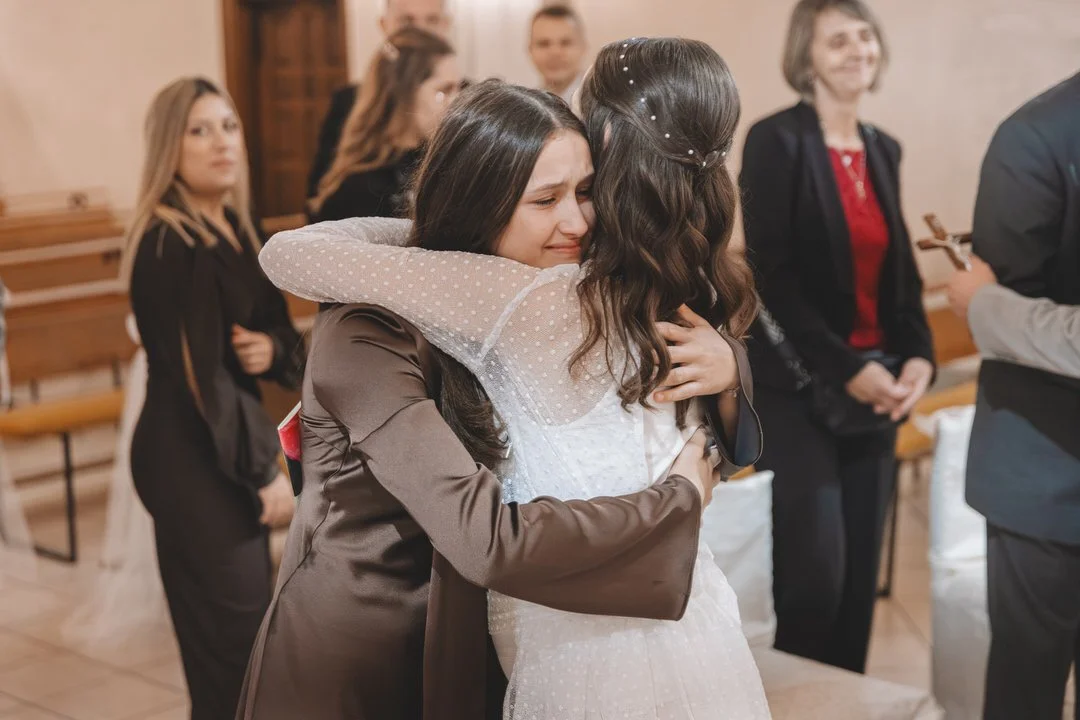 Two women hugging at a wedding ceremony, the bride and her sister sharing an emotional embrace while the sister is crying, with guests in the background.