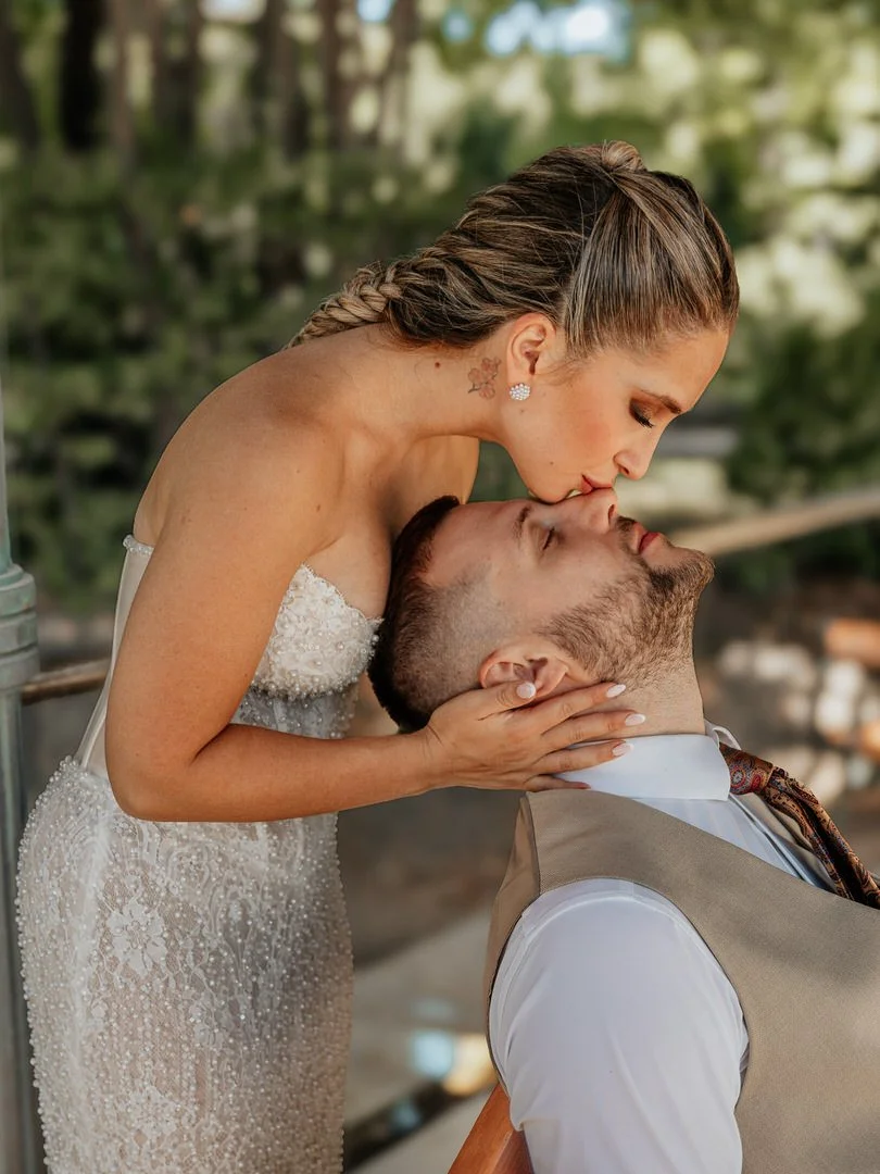 A woman in a white strapless dress leaning over a seated man, about to kiss him on the lips outdoors with trees in the background.