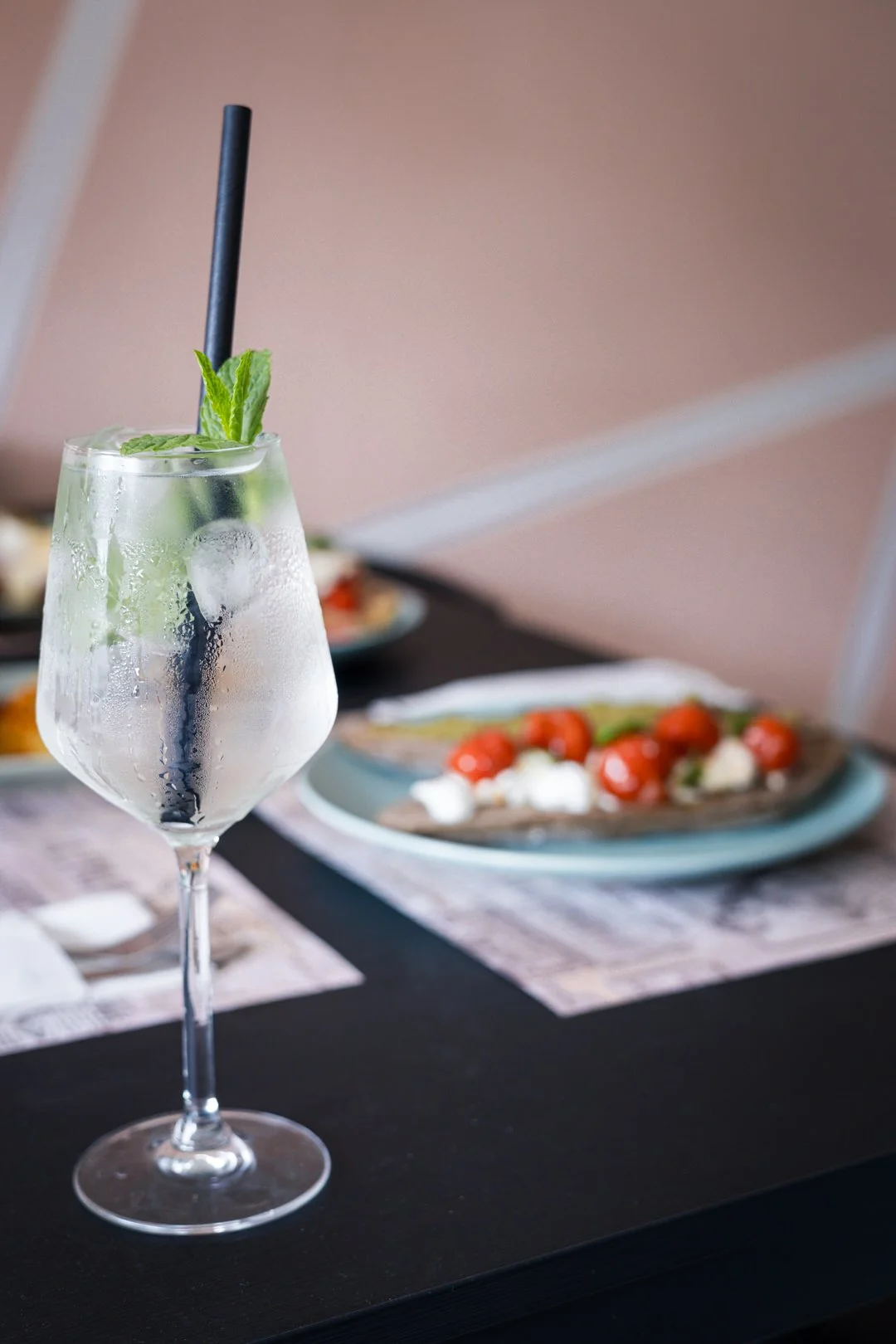 A glass of sparkling water with ice, lime slices, and mint leaves, placed on a dining table with plates of food in the background.