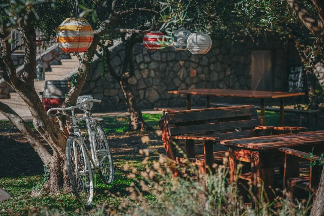 Outdoor seating area with wooden tables and benches surrounded by trees, hanging paper lanterns, a bicycle leaning against a tree, and stairs in the background.