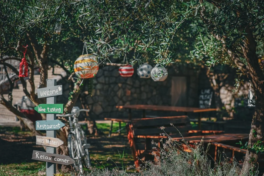 Outdoor garden with hanging ornaments, a wooden signpost pointing to locations such as welcome, wine tasting, restaurant, Halls kitchen, and relax corner, a bicycle leaning against a tree, and outdoor tables and benches in the background.