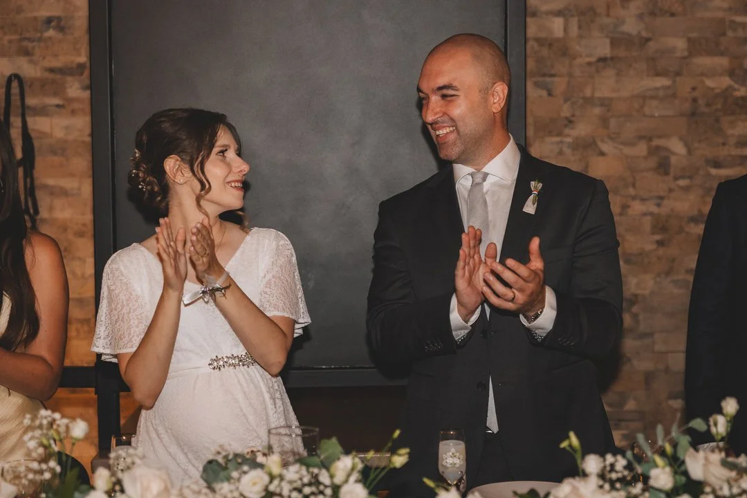 A man in a black suit and white tie is clapping and smiling at a woman in a white dress, who is also smiling. They are at a table decorated with flowers, likely during a celebration or wedding.